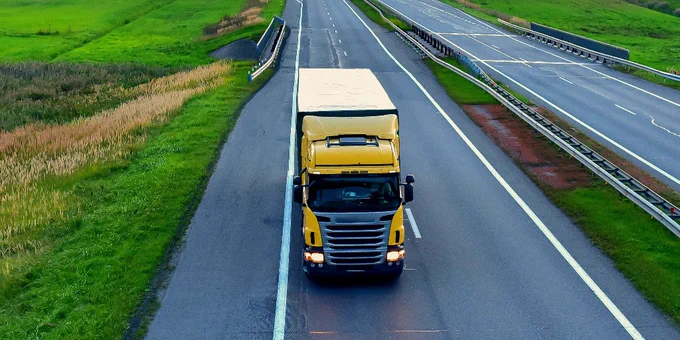 Aerial view of a yellow truck driving on a long, straight highway through a lush green landscape with fields on either side. The sky is clear with a warm, golden hue indicating either sunrise or sunset.