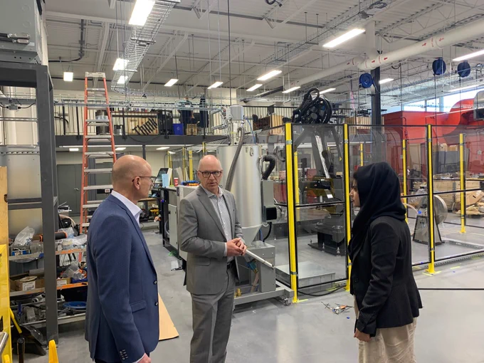 Alt text: Three people stand in conversation inside an advanced manufacturing lab filled with industrial equipment and machinery.