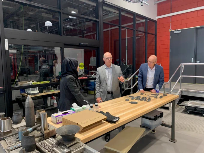 Alt text: Three people stand around a large worktable inside an advanced manufacturing and research lab. Two men in business attire and a student wearing a black headscarf discuss a collection of 3D-printed metal components and sample objects spread across the table.