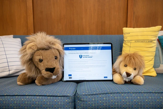 The lions Sol and Edgar are sitting on a blue couch at the State Library. In between them is an iPad and on the screen is the South Carolina Confederate Relic Room digital collection.
