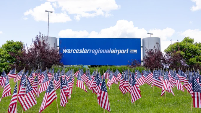 A field filled with numerous small American flags is in front of Worcester Regional Airport. The sky is partly cloudy, and trees surround the area.