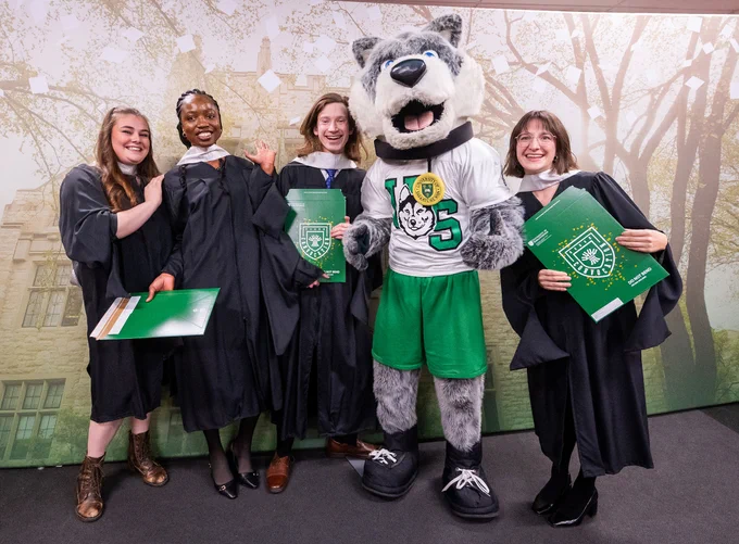A group of people in graduation robes stand smiling with a wolf mascot. They hold green University of Saskatchewan folders. The background features trees and a building façade.
