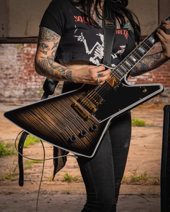 A person plays a Gibson electric Explorer guitar with a Figured top Cobra Burst finish. They have tattoos on their arms and are wearing a black shirt with a graphic design. Brick wall and concrete floor in the background.