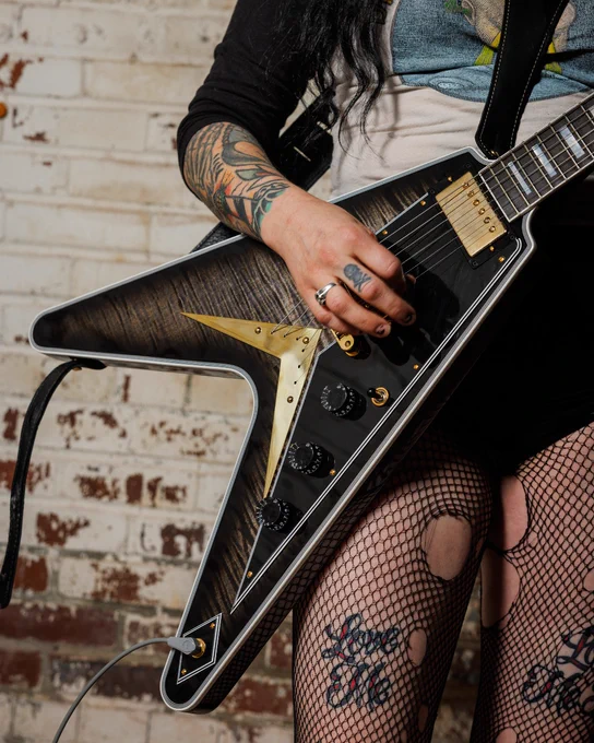 A person with tattoos is playing a black Gibson electric Flying V figured top, Cobra Burst guitar with gold accents, standing against a brick wall. They have fishnet leggings with the words "Love Me" visible.