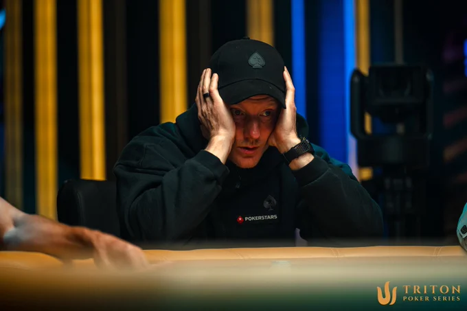 A focused poker player, wearing a black cap, rests his head in his hands, showcasing tension in a competitive setting.