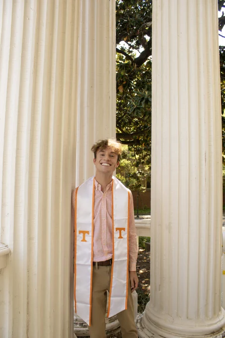 A person is smiling while standing between two large white columns. They are wearing a checkered shirt and a white stole with an orange letter "T" on each side. The background features foliage and sunlight filtering through.