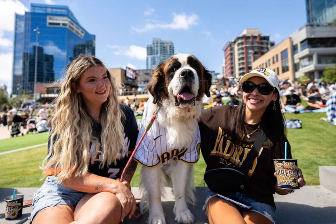 Photo of two Padres fans sitting next to their dog in Gallagher Square at Bark at the Park.