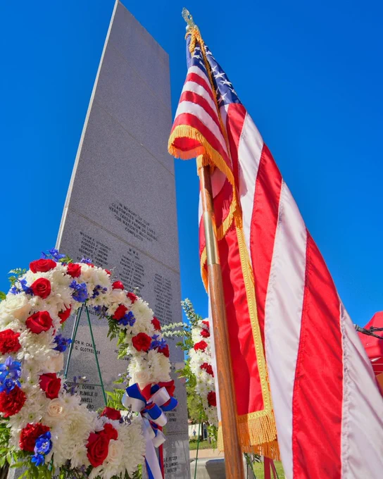 A tall stone memorial monument with engraved names stands against a clear blue sky. In front of it, an American flags is displayed, and a floral wreath with red, white, and blue flowers, adorned with a ribbon, is placed at the base.