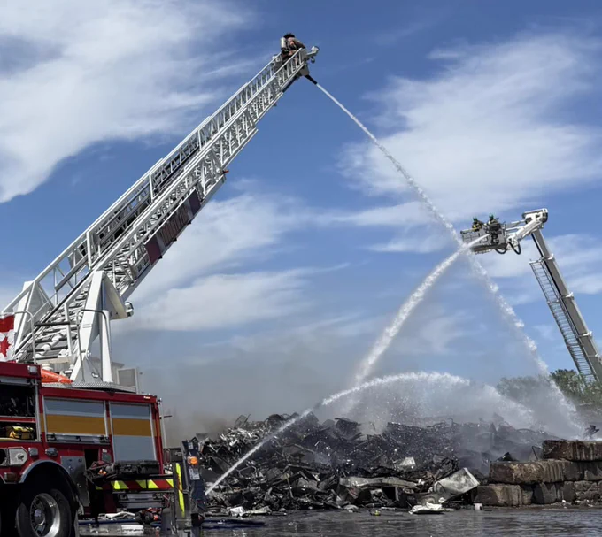 A rubble pile of smouldering materials being hosed down with water. On the left side of the photo is the rear end of a fire truck with an aerial ladder on the end spraying water. On the right side of the photo is another aerial ladder from a fire truck spraying water on the smouldering pile also.