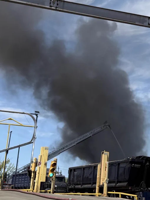 Black smoke plume covering the blue sky. In the forefront are trucks , stop light and hose and in the distance is an aerial ladder from a fire truck spraying water.