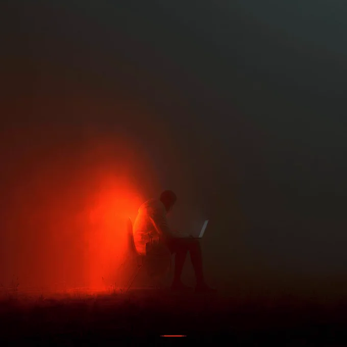 A figure sits in a chair, illuminated by a soft red glow, focused on a laptop in a dark, misty environment.