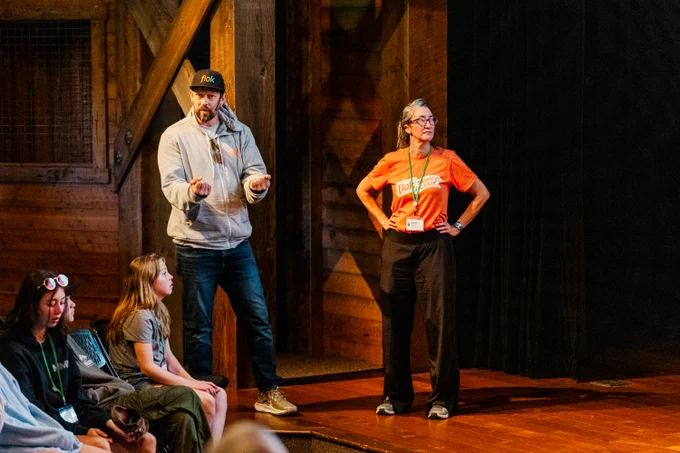 Two presenters stand on a wooden stage speaking to a small seated audience. One presenter gestures while speaking, and the other stands with hands on hips wearing a lanyard. Several attendees sit nearby, listening, in a warm indoor setting with wood-paneled walls.