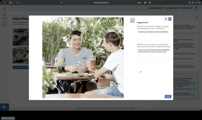 Two people sit at a small outdoor café table sharing a meal and conversation, with plates of food and drinks in front of them. One person has a visible prosthetic forearm. Green plants and soft natural light create a relaxed, social setting.