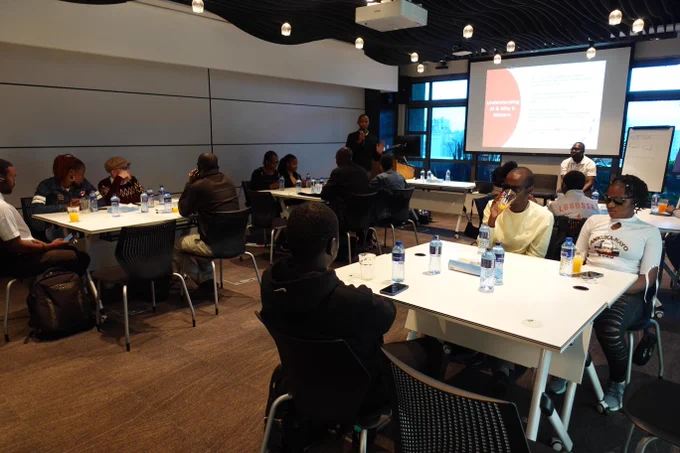 A group of people sit at tables in a modern meeting room while a presenter stands at the front speaking beside a projected slide that reads “Understanding AI & Why It Matters.” Attendees listen and take part in the discussion, with water bottles and notes on the tables.