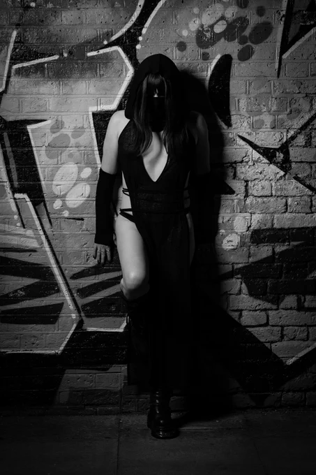 Trans Princess Jezebel is wearing a long flowing black cloak dress, long elbow length gloves and leather knee high boots as she leans against a wall covered in graffiti, as if laying in wait in the shadows [shot in black and white].
