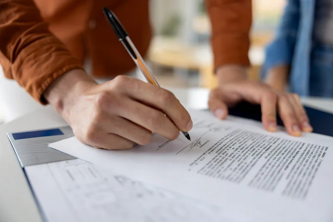 Close-up of a person signing a detailed contract document with a black pen on a clipboard.