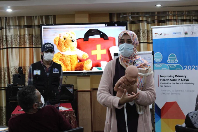 A masked Libyan woman demonstrates how to care for a baby (using a model) 