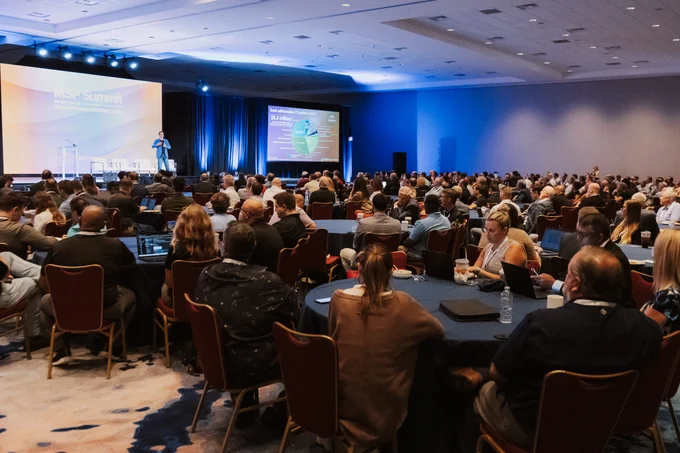 A keynote speaker on stage at MSP Summit showcasing their presentation in front a room of attendees who are watching the speaker on stage.