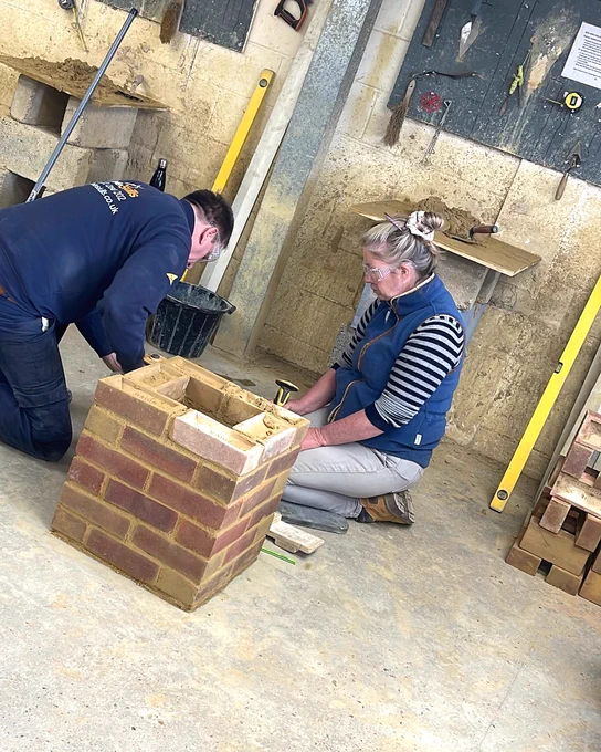 Two people working together on constructing a small brick structure indoors, surrounded by tools and construction materials.