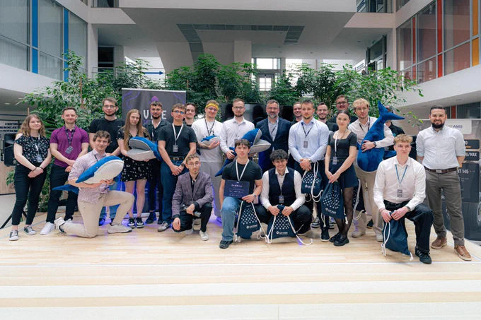 Uczestnicy wydarzenia Vibe the Future 2026 pozują do grupowego zdjęcia w przestrzeni Uniwersytetu Rzeszowskiego. Część osób trzyma pluszowe maskotki w kształcie wielorybów oraz torby z materiałami wydarzenia.