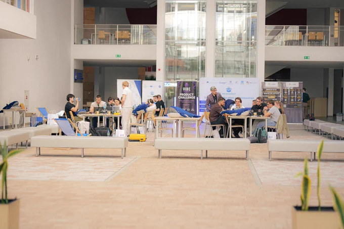 Przestrzeń wydarzenia Vibe the Future 2026 na Uniwersytecie Rzeszowskim. Uczestnicy pracują przy stołach, a w tle widoczne są roll-upy partnerów i materiały promocyjne wydarzenia.
