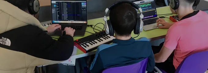 The backs of two young people and an adult sitting at a desk making music with keyboards, headphones and laptops.  