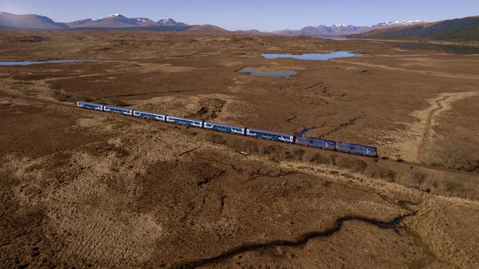 The Caledonian Sleeper travels through a vast, brown moorland with distant snow-capped mountains under a clear sky.