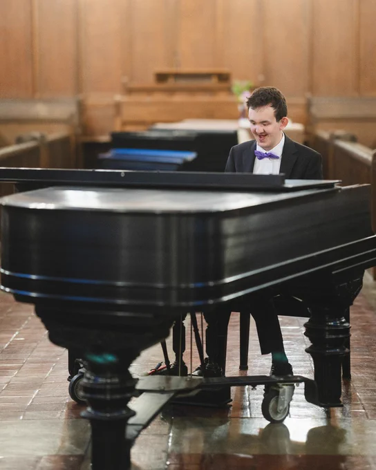 Matteo, wearing a black suit and purple bow tie, plays the piano in the chapel
