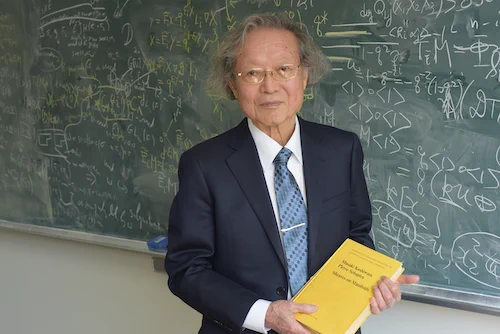 Masaki Kashiwara with one of his books next to a chalkboard covered with equations