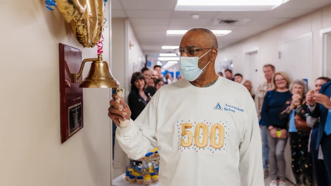 A person wearing a "500" shirt rings a bell in a hallway, marking a milestone. The hallway is lined with smiling onlookers and decorated with golden balloons.