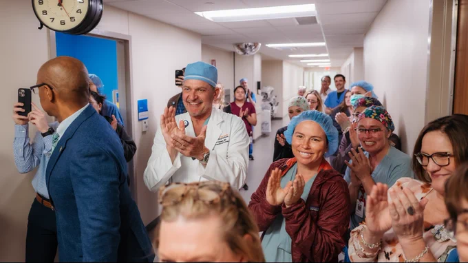 A group of medical professionals in hospital attire gather in a hallway, clapping and smiling.