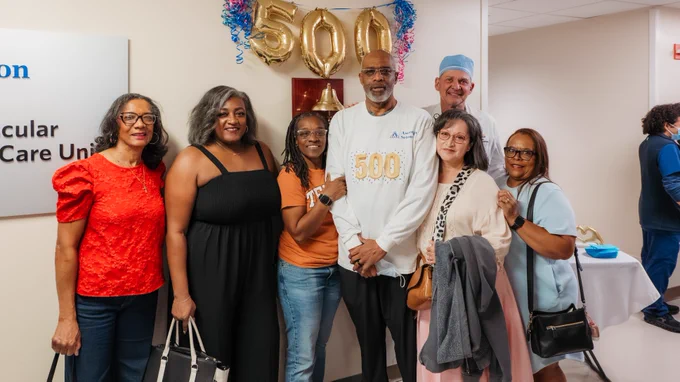 A group of people stands together in a hospital setting. One person is wearing a shirt with "500" printed on it, matching the "500" balloons behind them. They are smiling and posing for the photo. A medical professional is in the background.
