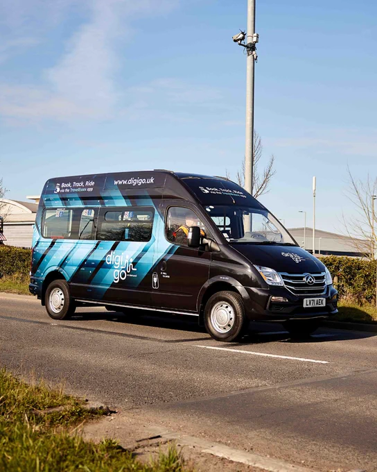 A black 'DigiGo' minibus with blue graphics driving on a road. Book, Track, Ride via the TravelEssex app.
