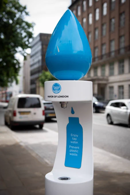 A public drinking water refill station on a London street, topped with a large blue droplet. The unit displays Thames Water and Mayor of London branding, with a blue bottle graphic and text reading “Enjoy tap water. Prevent plastic waste,” with parked cars and buildings visible in the background.