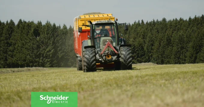 Ein Traktor ist frontal auf einem Feld mit gem\u00E4htem Gras zu sehen, im Hintergrund ist ein dichter Wald zu erkennen. An den Traktor ist ein rot-gelber Anh\u00E4nger angekuppelt.