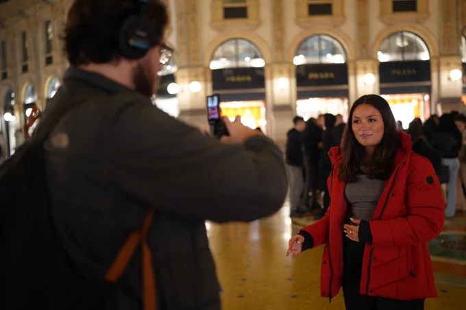 A woman in a red coat stands indoors, facing a man with headphones filming her. The elegant, warmly-lit setting is filled with people in the background.