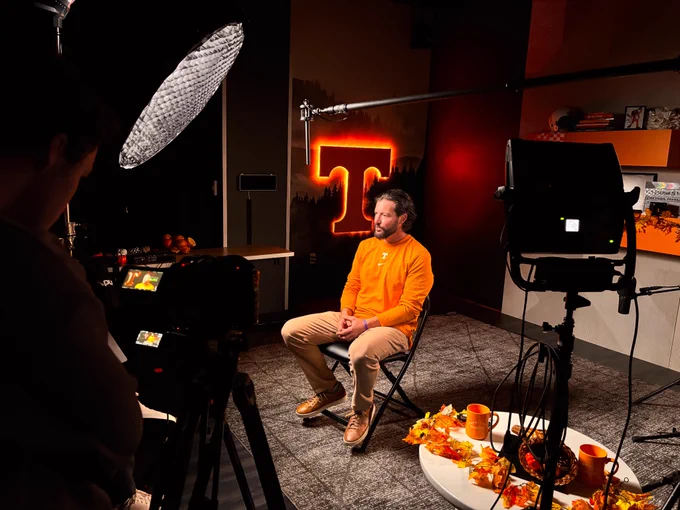 A person in an orange shirt is seated in a dimly lit room with a glowing orange "T" on the wall behind. Camera equipment surrounds the scene, and a table with autumn leaves and mugs adds a warm, focused atmosphere.