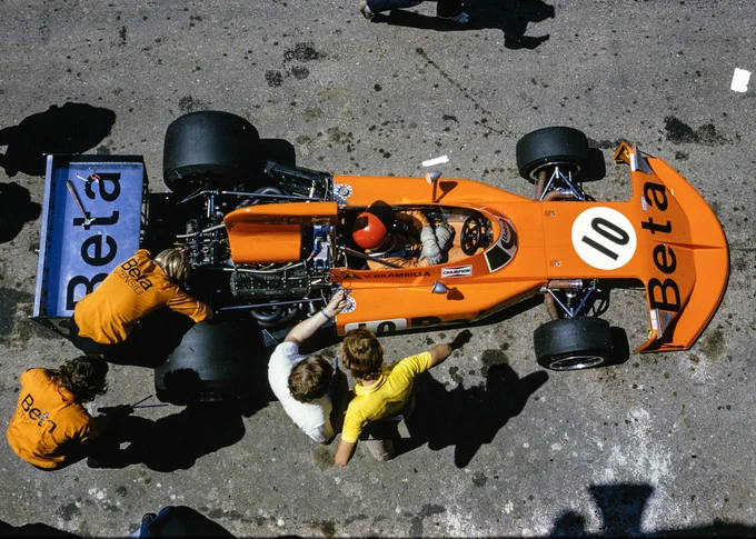 A high angle view, almost aerial, of the Beta sponsored March 741 in pit lane, facing rightwards. The car is orange with a powder blue rear wing, and has Vittorio Brambilla at the wheel, wearing an orange race suit, grubby white gloves and a red crash helmet. March mechanics work on the back of the car as other team members are in conversation.