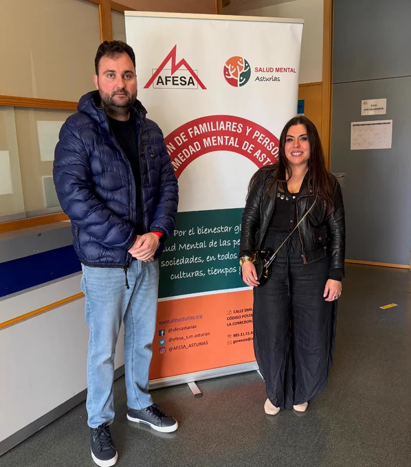 Dos personas posan en el interior de un edificio junto a un roll-up de AFESA Salud Mental Asturias. El cartel incluye el logotipo de la entidad y el texto sobre la asociación de familiares y personas con enfermedad mental. La escena se desarrolla en un espacio interior con paredes claras, puerta al fondo y suelo gris.