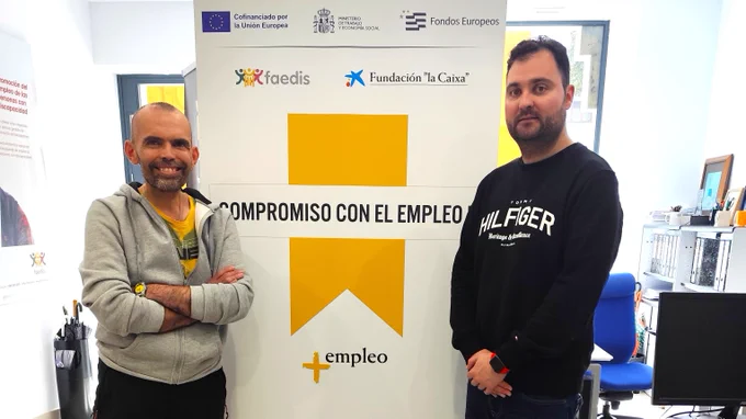 Dos personas posan en una oficina delante de un roll-up de FAEDIS con el texto “Compromiso con el empleo”, acompañado de logotipos de entidades colaboradoras como Fundación “la Caixa” y el Fondo Social Europeo. Alrededor se observan mesas de trabajo, una silla de oficina y material informático.