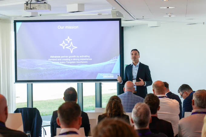 Business professional presenting a mission statement to an attentive audience in a modern conference room with a large screen displaying key goals.