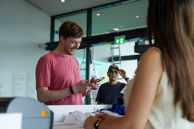 A man in a pink shirt uses his phone while standing at a counter with a woman facing him in a modern indoor setting.