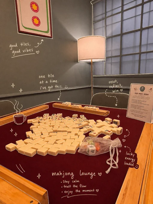 A warm editorial-style photograph inside Mahjong Lounge in Koramangala, Bangalore featuring a premium Mahjong table with cream Mahjong tiles scattered across a deep burgundy playing surface. Soft ambient lighting from a standing floor lamp creates a calm and cozy atmosphere. Hand-drawn white doodles and handwritten-style text overlays surround the scene with phrases like “good tiles, good vibes,” “one tile at a time,” and “reset, reflect, win.” A small illustrated coffee cup doodle adds to the relaxed café aesthetic. Wooden Mahjong racks frame the table while Mahjong tiles with traditional Chinese characters are visible throughout the image. The composition reflects the thoughtful and mindful experience of playing Mahjong at Mahjong Lounge, highlighting strategy, social connection, community gaming, and slow evenings in a beautifully curated lounge setting in Bangalore.