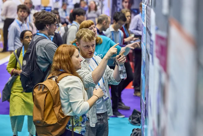 Group of people attentively examining and pointing at information displayed on a large board at a busy event or conference.