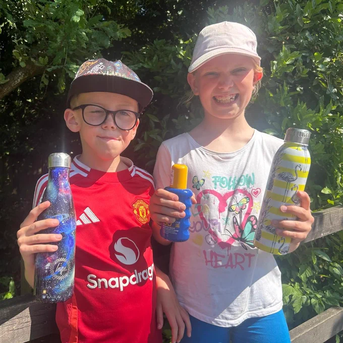 Two children holding water bottles and sunscreen, smiling at the camera.