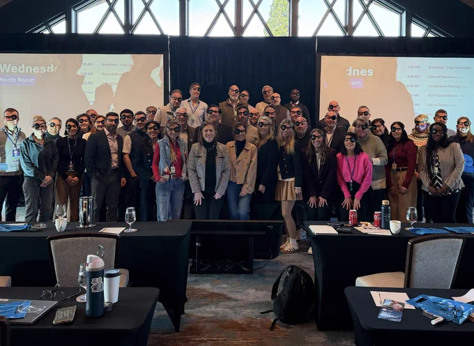 Delcath team members pose together at a company meeting wearing eye patches to raise awareness for ocular melanoma and Eye Patch Day. The group stands in front of presentation screens inside a conference venue.