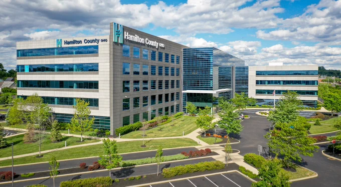 A modern office building with reflective windows and Hamilton County, Ohio signage, surrounded by landscaped greenery and parking lots under a partly cloudy sky.