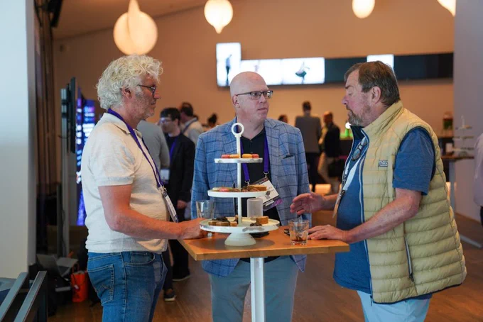 Three men engaged in conversation around a high table with a tiered tray of assorted desserts in a modern event space.