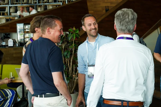 Group of men engaged in conversation at a professional event in a modern indoor setting with wooden walls and green seating.