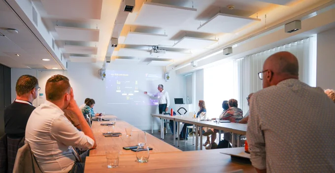 A presenter explains data on a whiteboard to a group seated around a conference table in a modern meeting room.